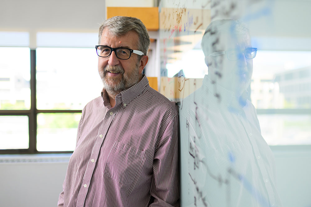 Faculty member Curtis Smith leaning on a glass whiteboard with writing inside and office with a window in the background