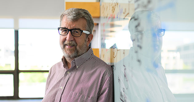 Faculty member Curtis Smith leaning on a glass whiteboard with writing inside and office with a window in the background, MIT