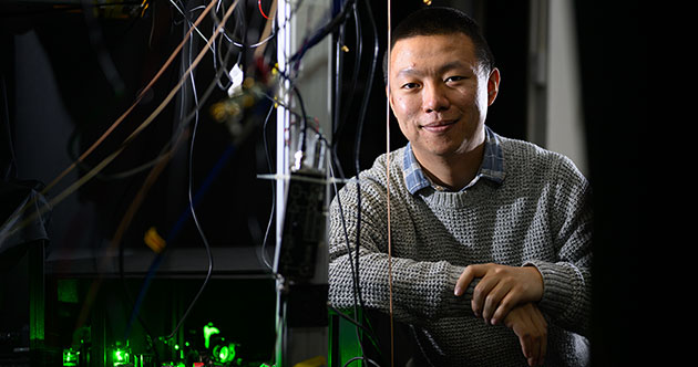 Male postdoc, Guoqing Wang, in lab leaning on his right arm; green laser instrumentation set-up in the foreground to the left and wires hanging above, MIT