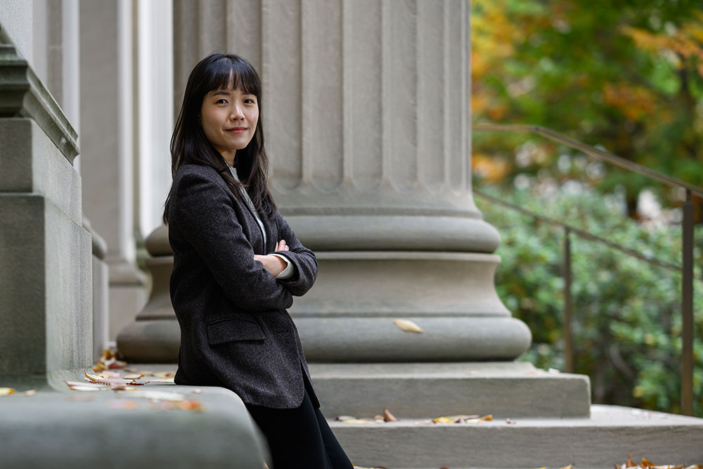 Female grad student Youyeon Choi, arms folded outside on a path with trees on the right.