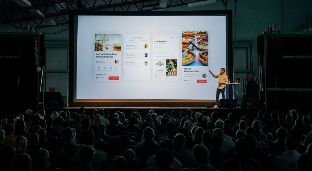 person discussing while standing in front of a large screen in front of people inside dim-lighted room